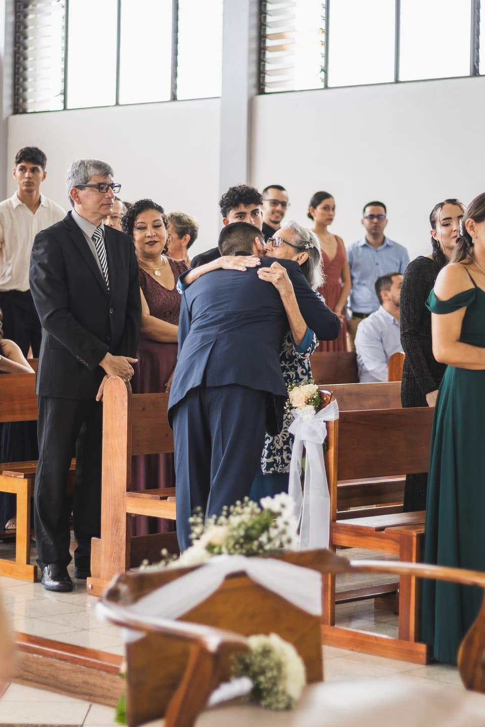 Dos personas se abrazan con emoción dentro de la iglesia, rodeadas de invitados