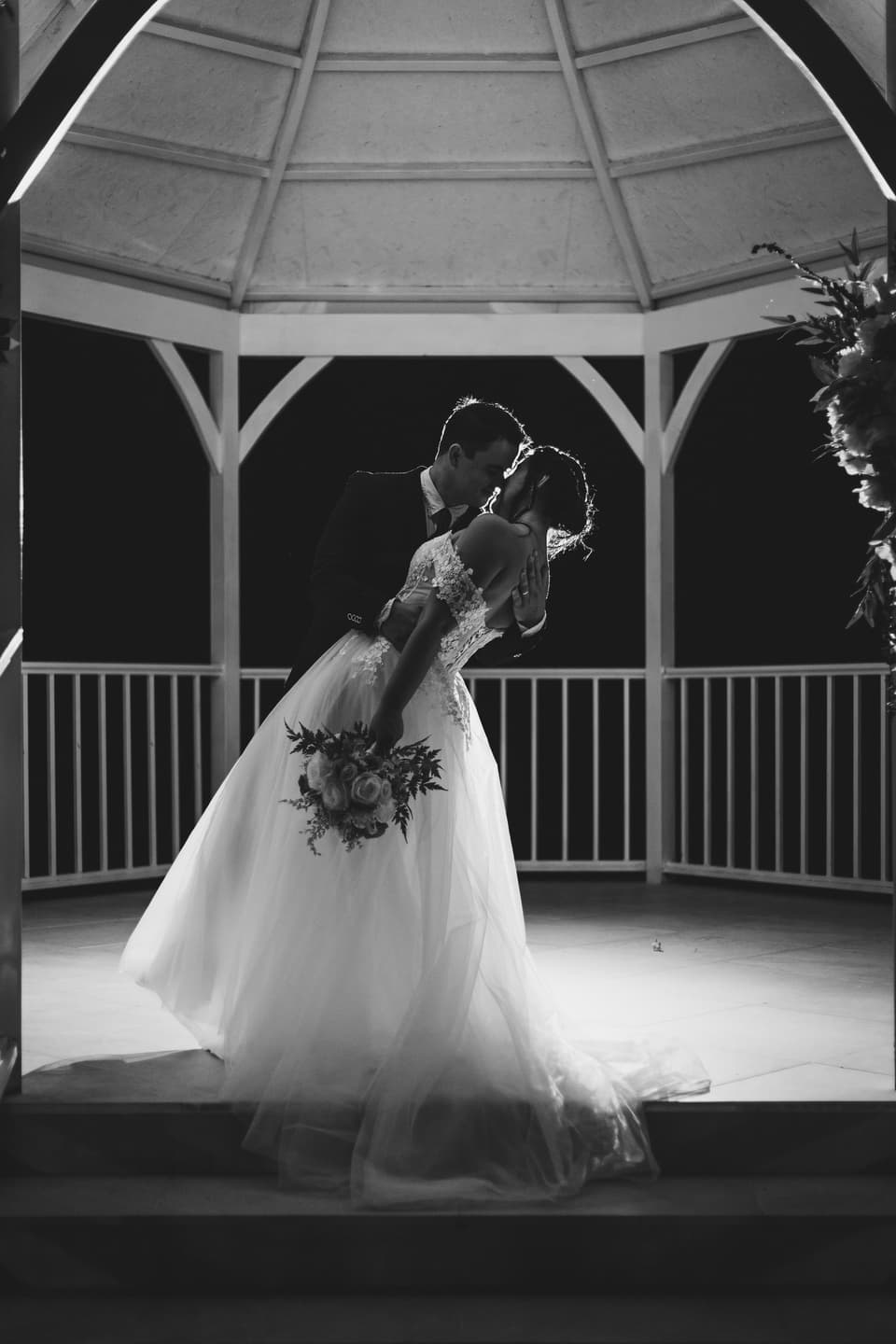 Beso inclinado de los novios dentro de un gazebo, en siluetas y blanco y negro
