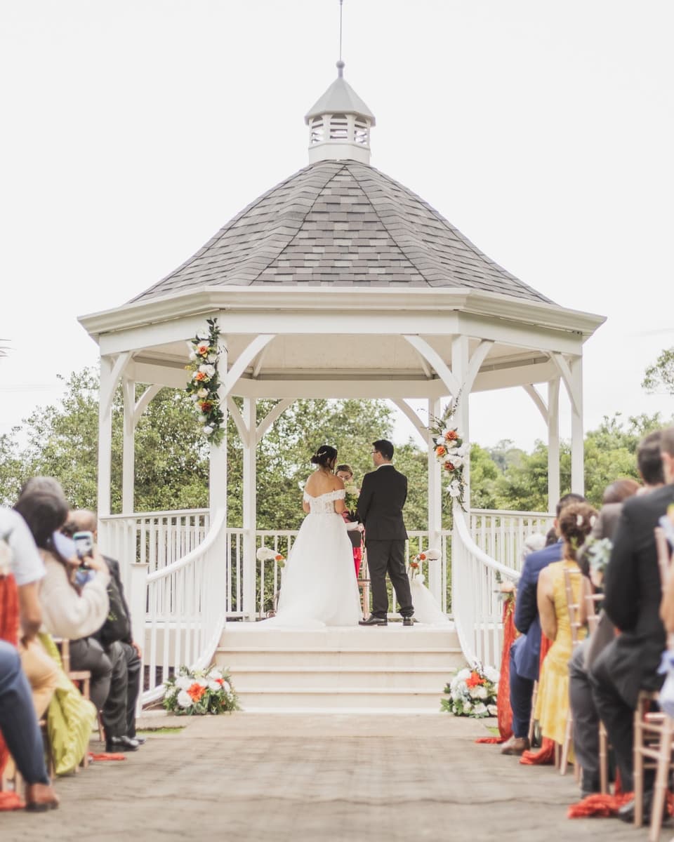 Ceremonia al aire libre en un gazebo blanco rodeado de palmeras, con los invitados sentados