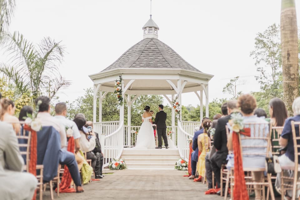 Ceremonia al aire libre en un gazebo blanco rodeado de palmeras, con los invitados sentados