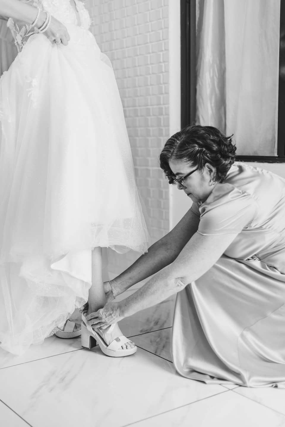 Una mujer mayor agachada ayudando a la novia a ponerse los zapatos, en blanco y negro