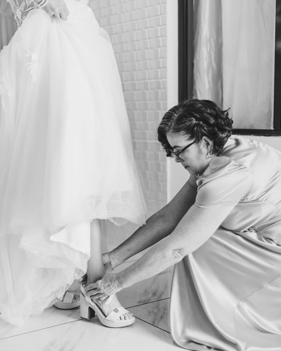Una mujer mayor agachada ayudando a la novia a ponerse los zapatos, en blanco y negro