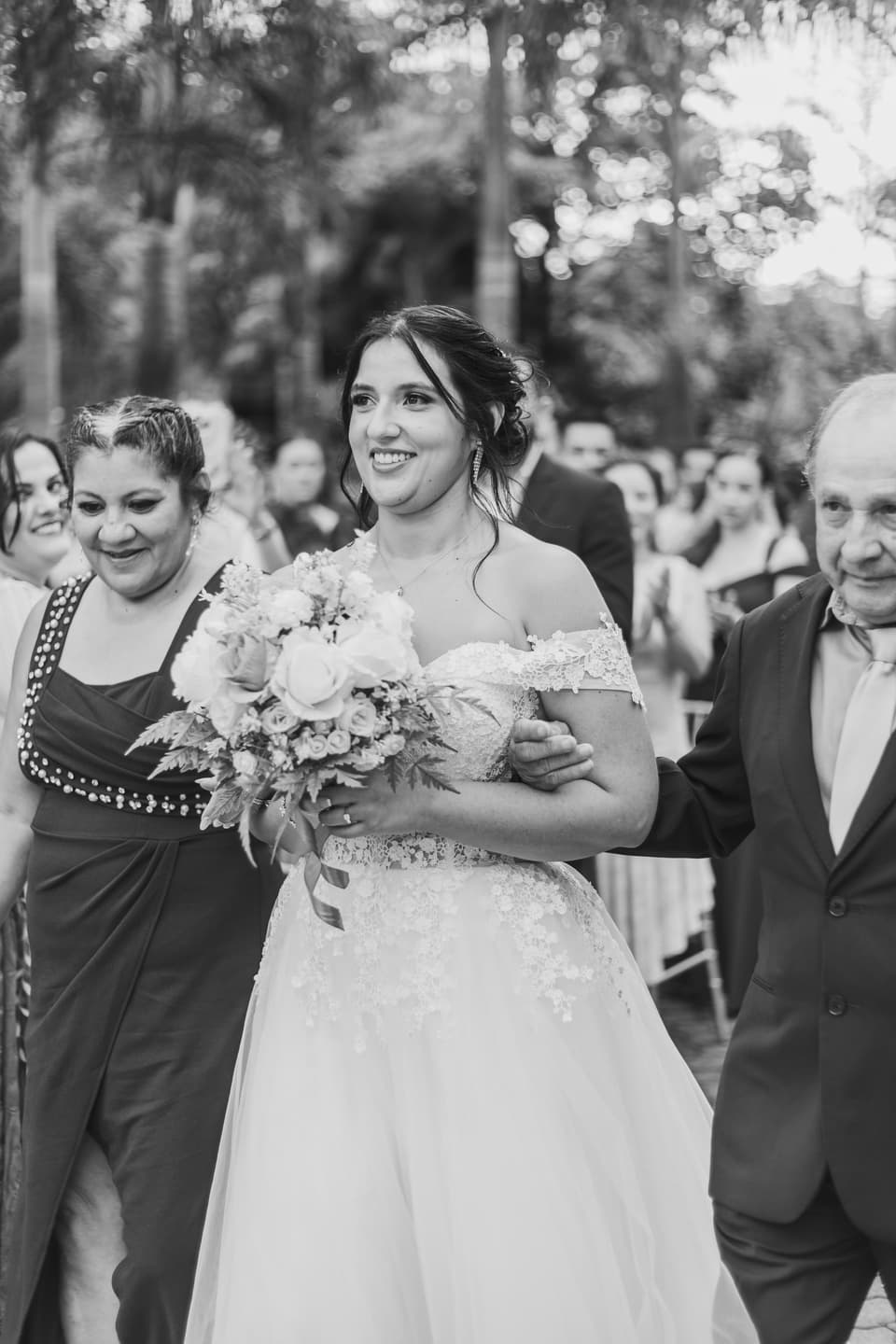 Novia sonriendo con su ramo mientras camina al altar acompañada de dos personas, en blanco y negro