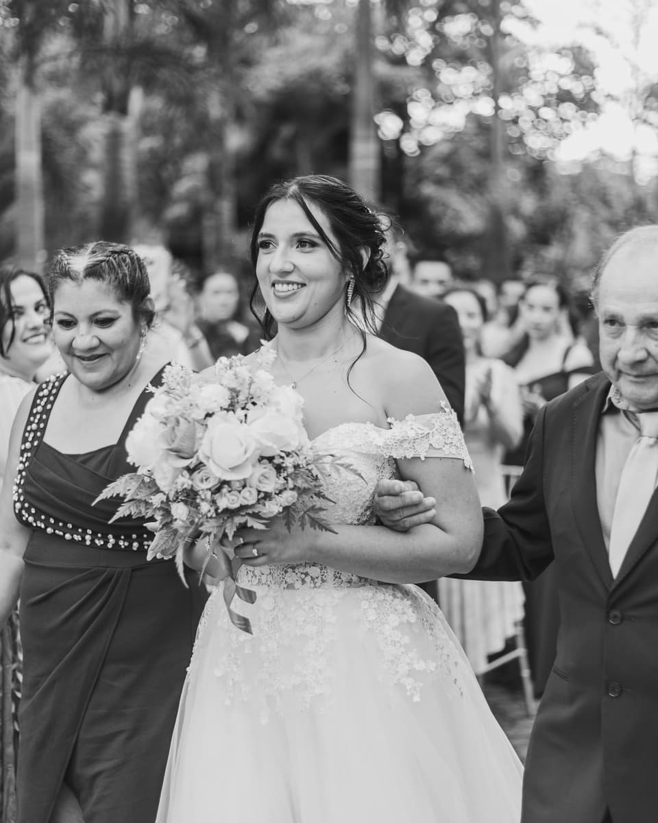 Novia sonriendo con su ramo mientras camina al altar acompañada de dos personas, en blanco y negro