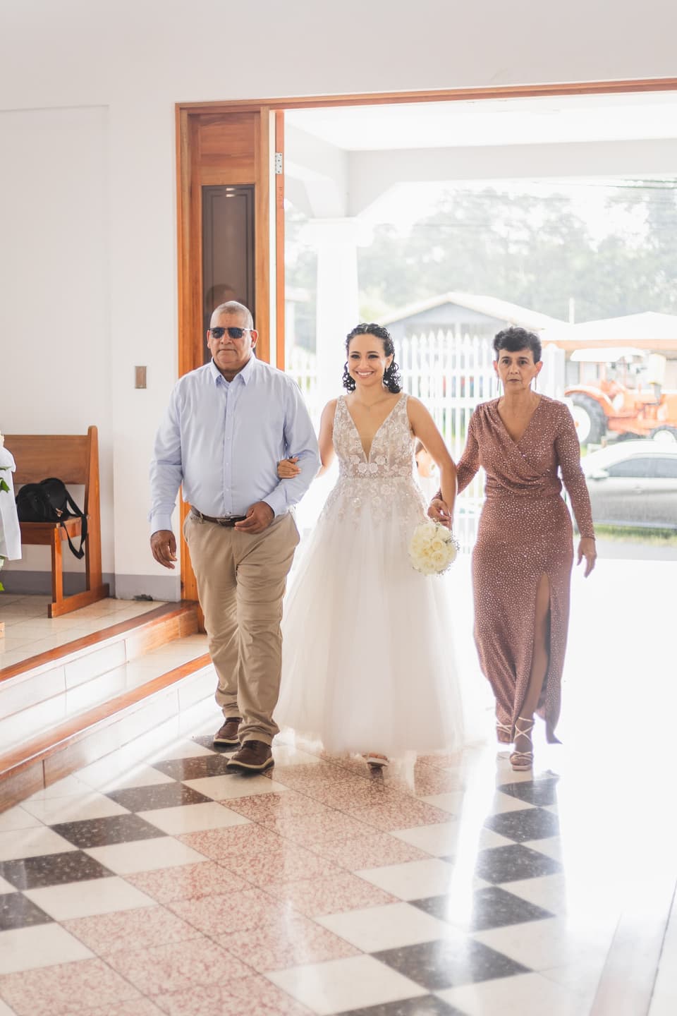 Novia entrando a la iglesia entre un señor y una señora que la acompañan