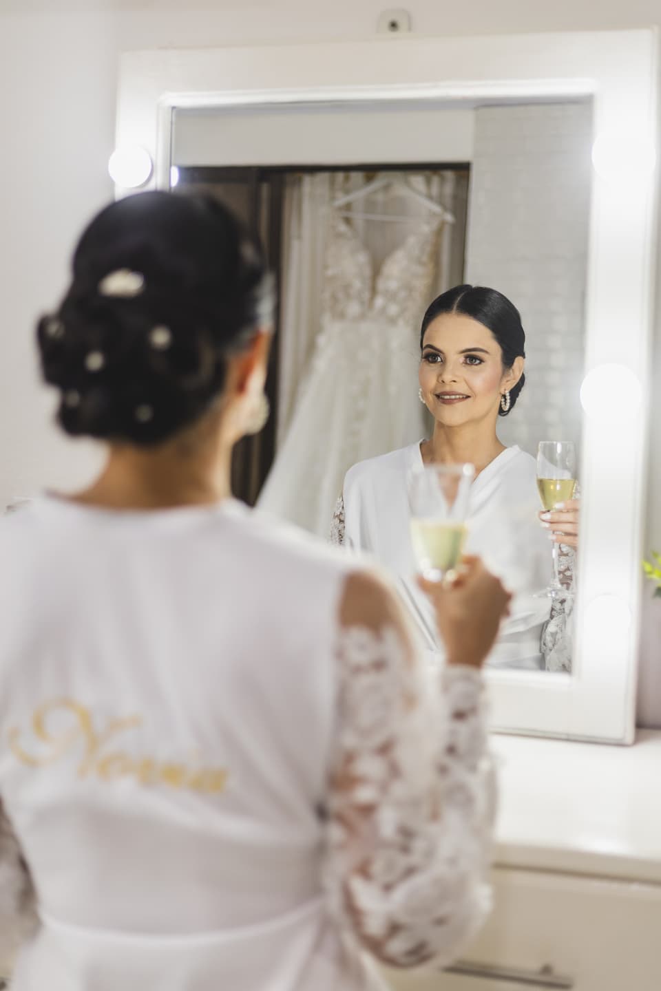 Novia frente al espejo con una copa en la mano, con su vestido colgado detrás