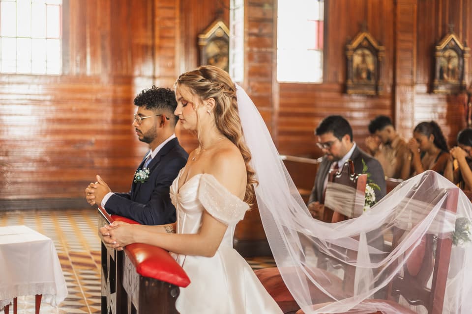 Novios arrodillados frente al altar en una iglesia de madera, con el velo largo en el aire