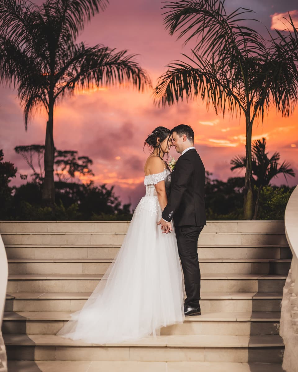 Novios con las frentes juntas sobre unas gradas, con palmeras y un cielo rosado al atardecer