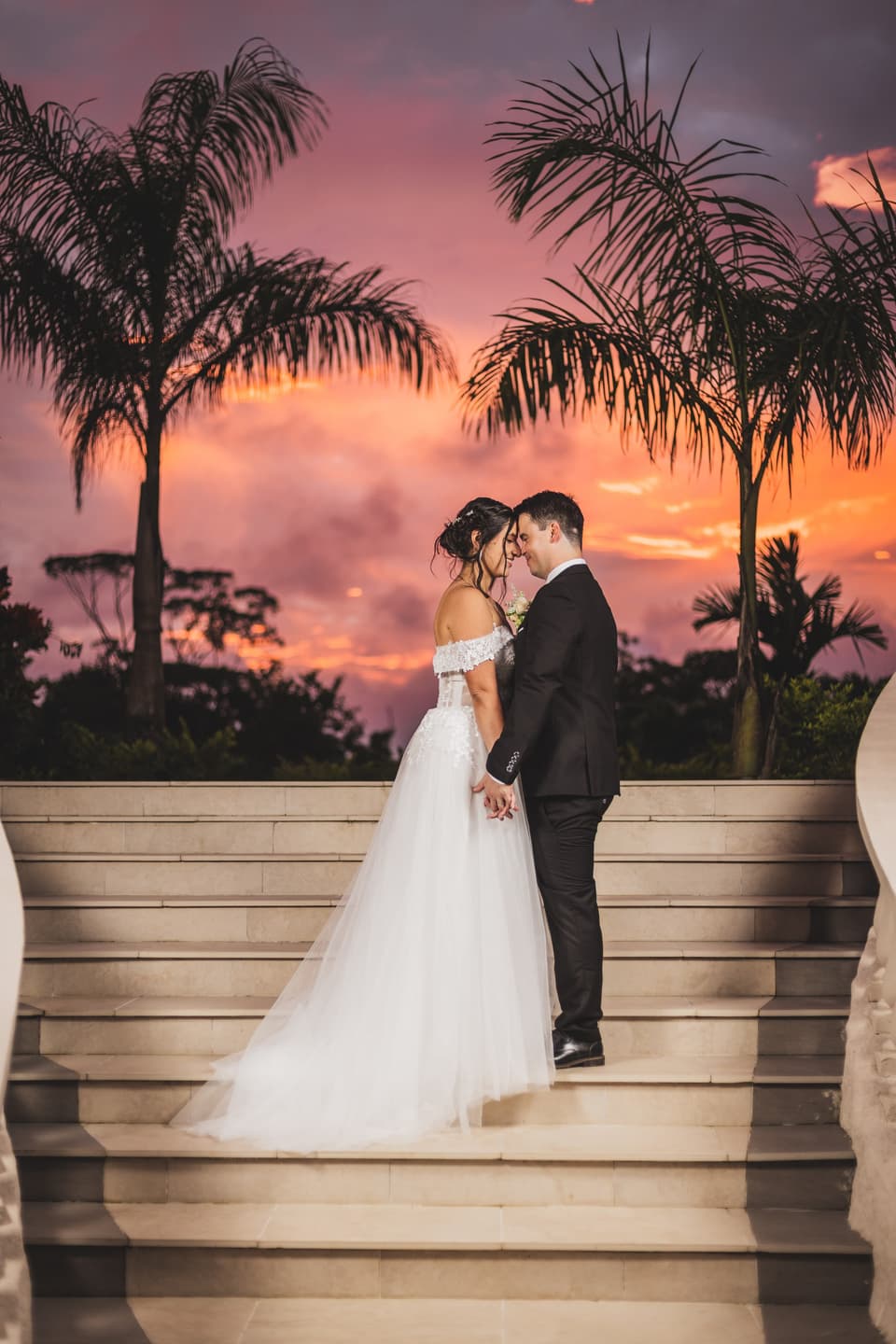 Novios con las frentes juntas sobre unas gradas, con palmeras y un cielo rosado al atardecer