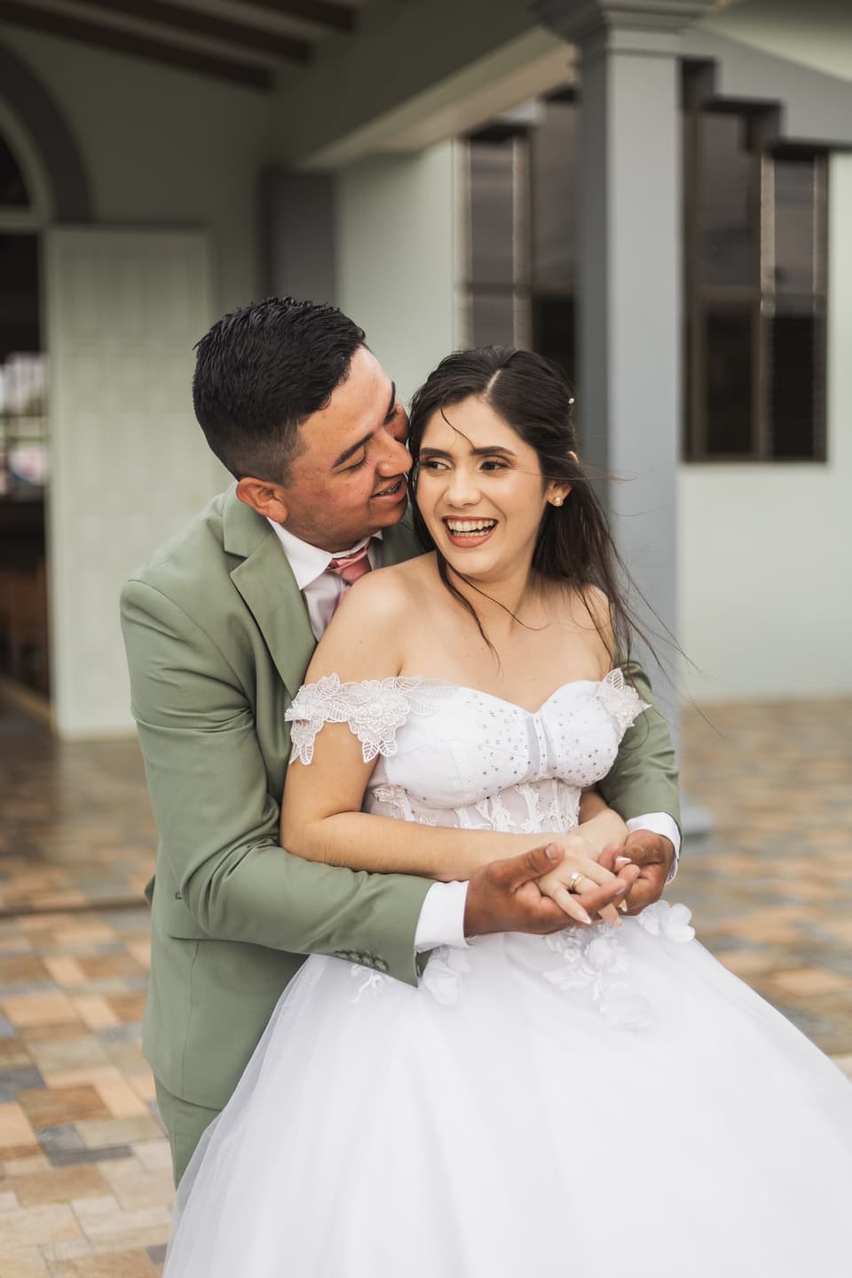 Novios abrazados, ella riendo con ganas, frente a una casa de corredor