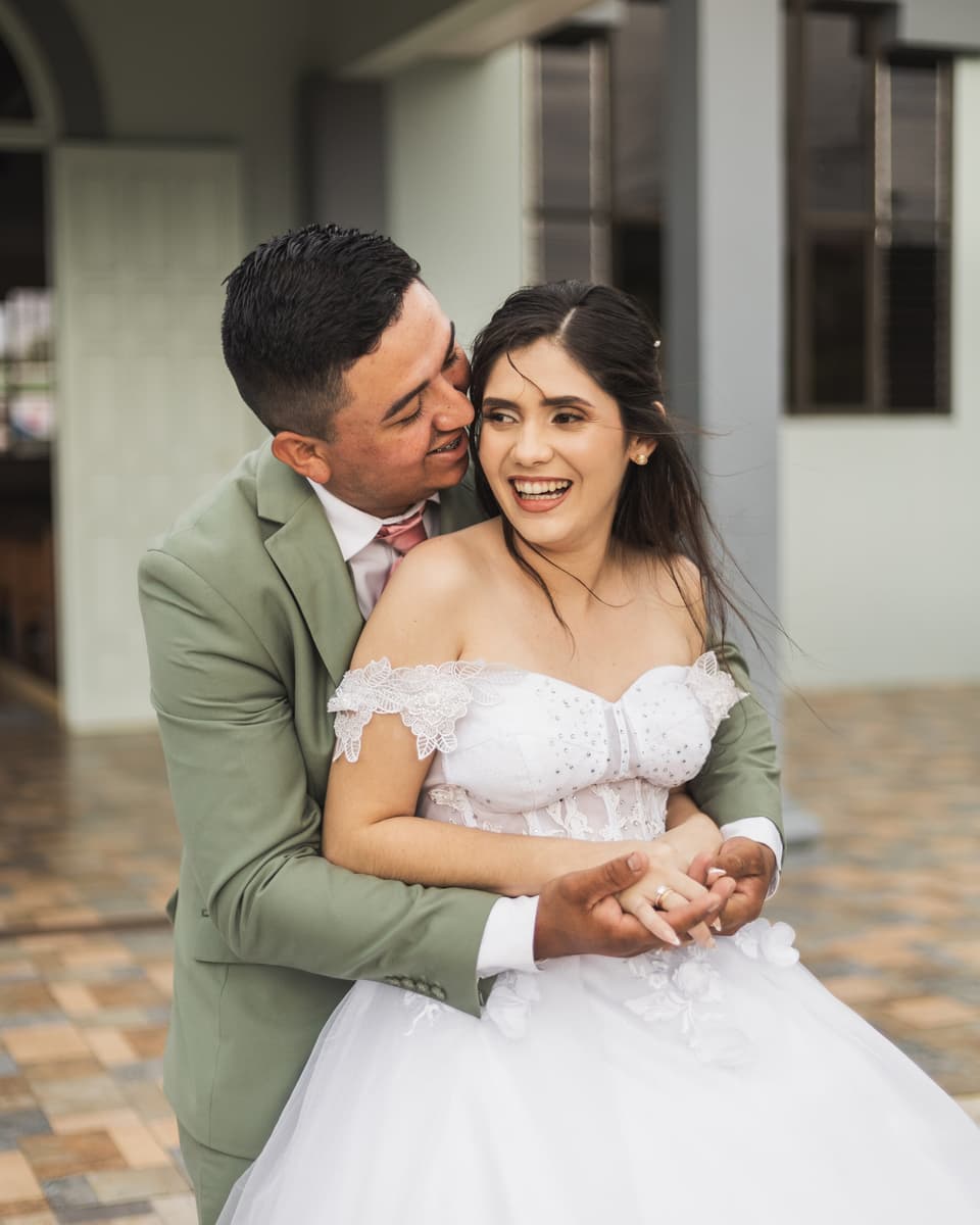 Novios abrazados, ella riendo con ganas, frente a una casa de corredor