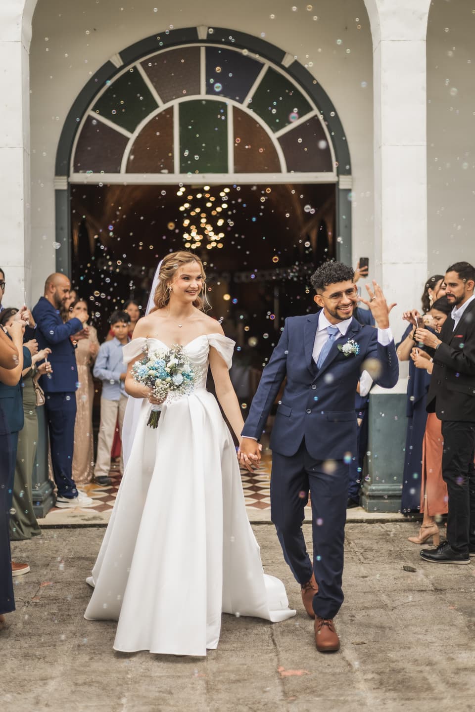 Novios saliendo de la iglesia sonriendo, entre invitados y burbujas