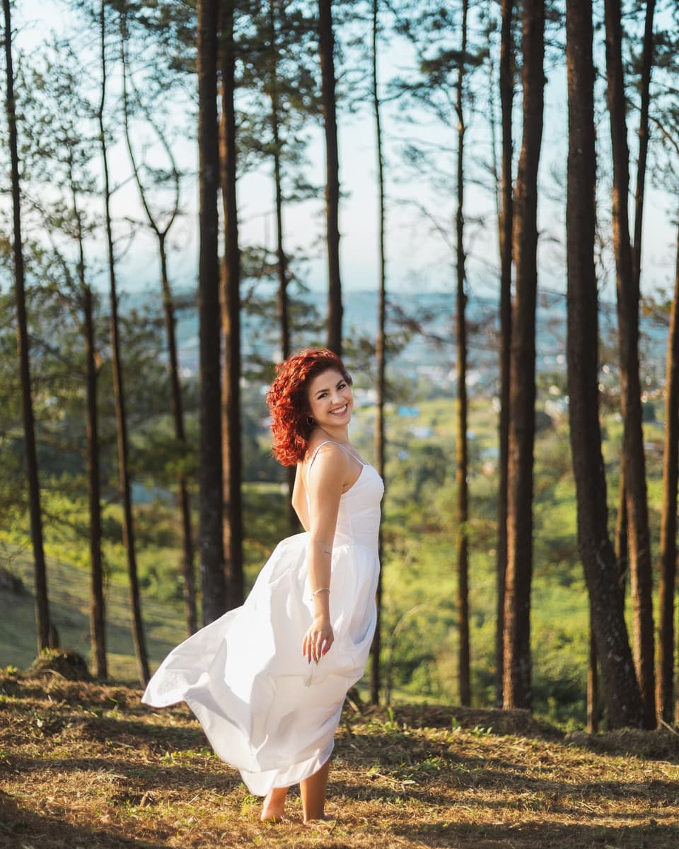 Fabiola Soto en un bosque de pinos, con el vestido blanco moviéndose y el valle al fondo