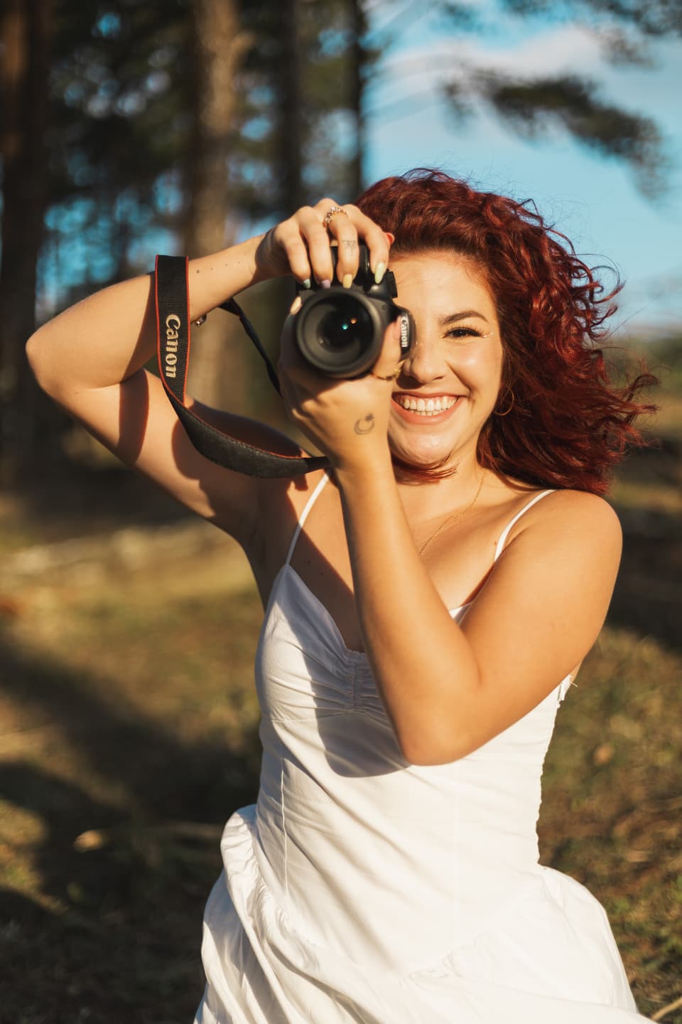 Fabiola Soto sonriendo mientras toma una foto con su cámara, con luz de tarde entre los árboles